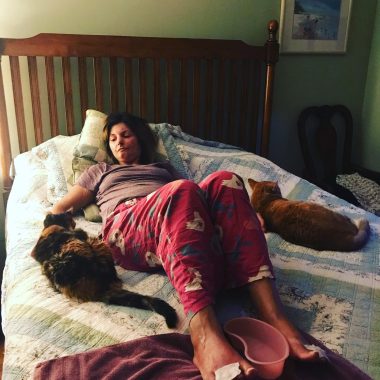 living with scleroderma | Scleroderma News | photo of Amy lying on her bed, in red pajama bottoms and a gray T-shirt, with a cat on either side of her