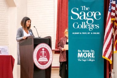comprehensive care | Scleroderma News | Amy stands behind a lectern that has a college seal on the front. To her side is a green flat with the words "The Sage Colleges" in large type, with a United States flag beside it. 