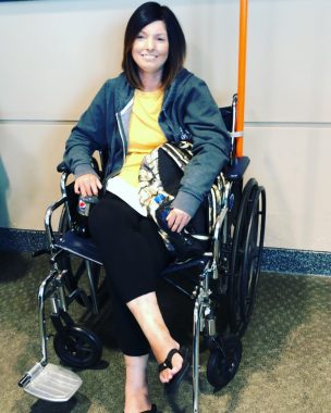 Amy smiles while seated in her wheelchair at the airport. She's wearing a yellow shirt, dark jacket, black leggings, and flip flops.