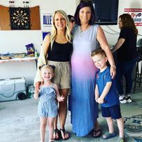 Two women stand side by side with their arms around each other. The woman on the left, Kelly, has blonde hair and is wearing a black tank top, tan shorts, and black sandals. The woman on the right, Amy, has brown hair and is wearing a pink and blue maxi dress and black sandals. Kelly's young son and daughter stand on either side of the women as they all pose in what appears to be a garage.