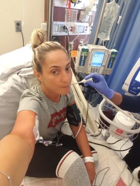 A blonde-haired woman is in a hospital bed with machines and tubes around her. A blue-gloved hand is taking her temperature. She wears a gray T-shirt with what appears to be a red logo, as well as black pants with long, gray and white socks. One of her arms is outstretched toward the camera; the other wears a blood pressure monitor. 