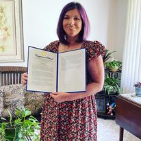 A woman with shoulder-length purple hair stands in what appears to be a living room holding a two-page letter mounted on a blue background. She's wearing a long floral dress, and there are several plants and pieces of furniture around her.