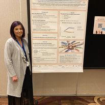 A woman stands next to a large abstract poster at a scleroderma conference. She has dark shoulder-length hair and is wearing a black dress, long gray cardigan, and a blue lanyard. We can't see what's on the poster, but there appear to be several graphics amid the text.